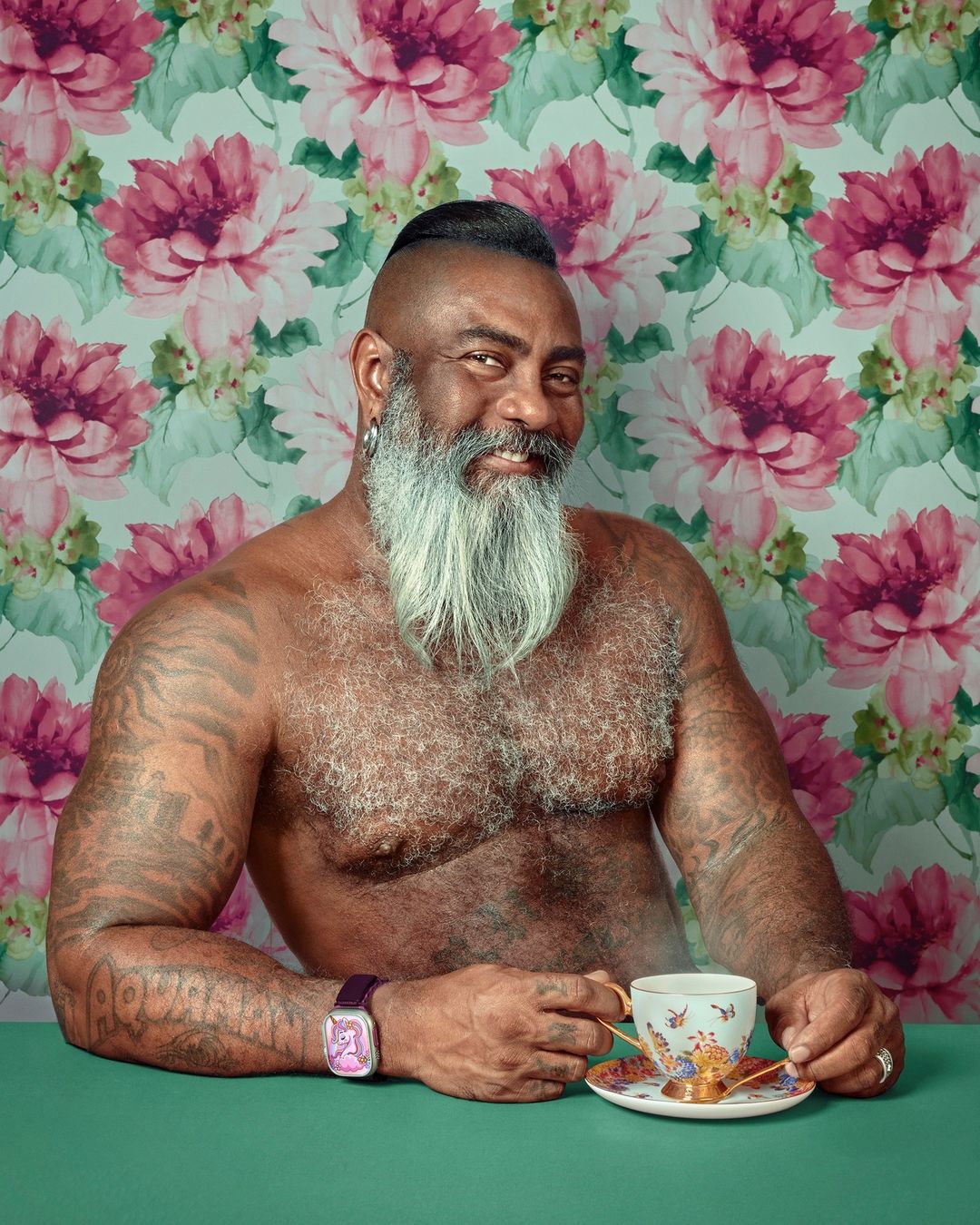 Portrait of a mature adult male with tattoos, a beard, and an unusual haircut drinking tea, against a floral wallpaper backdrop.